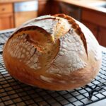 A freshly baked gluten-free sourdough loaf with a golden, crispy crust sitting on a cooling rack. The bread looks rustic with natural cracks on the crust, and there’s a warm kitchen in the background.