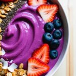 A vibrant smoothie bowl with a thick, purple base made from blended bananas and berries, topped with gluten-free granola, chia seeds, sliced strawberries, blueberries, and coconut flakes.